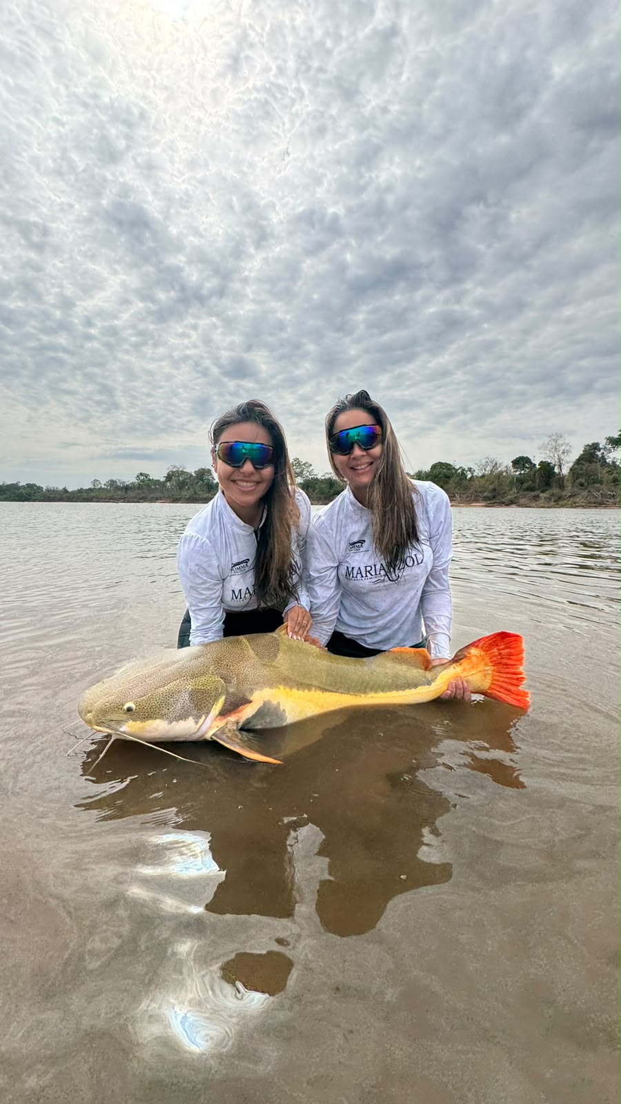 Ana Claudia e Anna Hiria juntas no rio com um peixe, usando camisetas da Marianzol